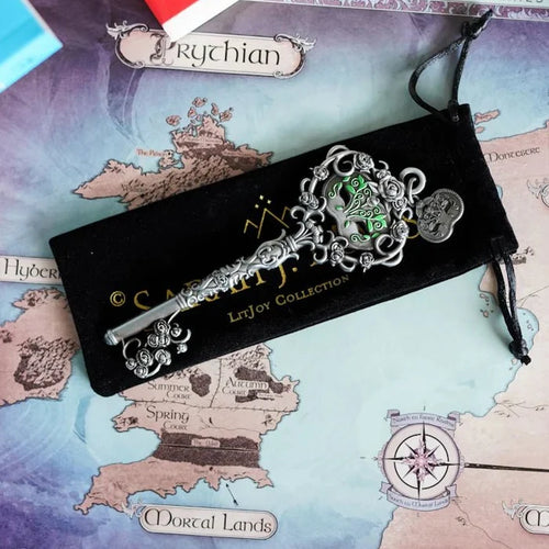 Officially licensed ACOTAR Spring Court key by Sarah J. Maas LitJoy Collection — ornate silver key with green enamel floral bow and rose details, resting on its black velvet drawstring pouch on the Prythian map, with ACOTAR series books in the background