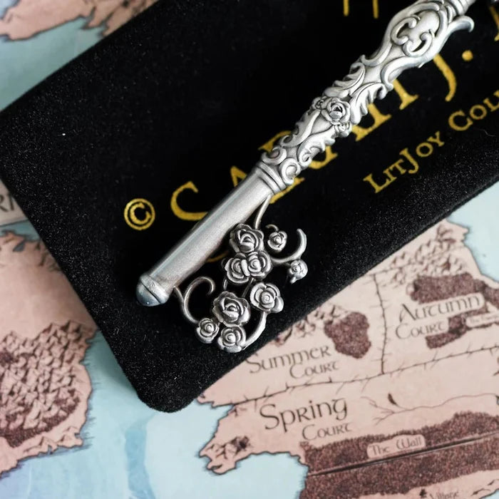 Close-up of the officially licensed ACOTAR Spring Court key shaft and bit by Sarah J. Maas LitJoy Collection — silver key with a rose bouquet-shaped key bit and intricate floral scrollwork shaft, on the branded velvet pouch, over the Prythian map highlighting Spring Court, Summer Court, and Autumn Court