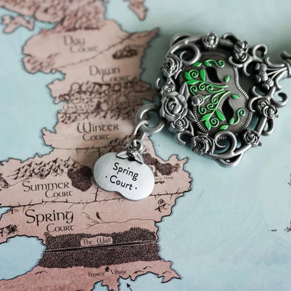 Close-up of the officially licensed ACOTAR Spring Court key bow by Sarah J. Maas LitJoy Collection — silver rose filigree frame with glowing green enamel vine centerpiece and engraved oval "Spring Court" hanging tag, positioned directly over the Spring Court territory on the Prythian map