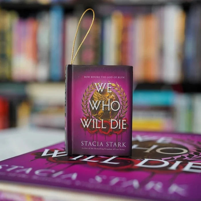 We Who Will Die miniature book ornament by Stacia Stark — tiny purple hardcover with gold laurel wreath and gold loop hanger, standing on the full-size edition with a colorful bookshelf blurred in the background