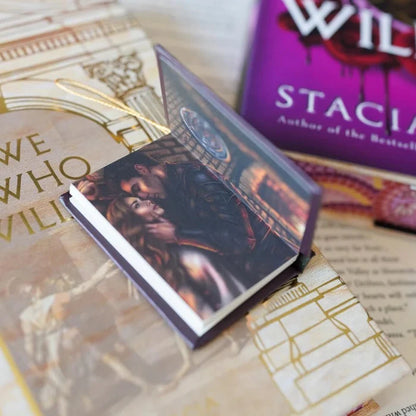 We Who Will Die LitJoy mini book ornament opened to a full-color interior illustration of a red-haired woman in a dramatic architectural scene, resting on the beige gold LitJoy exclusive cover with the purple edition in the background